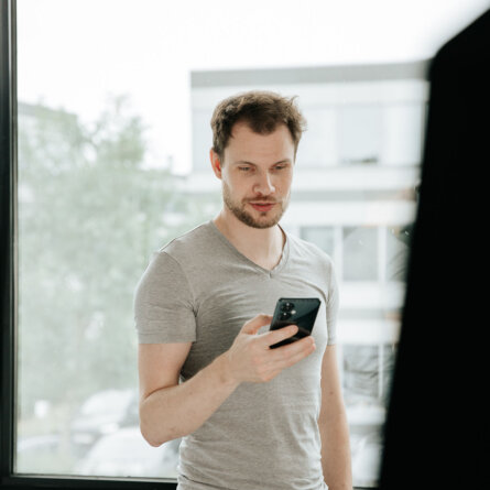 A man in a light grey t-shirt stands by a large window, holding a smartphone and looking at the screen with focus. The background shows a blurred office building and trees outside.