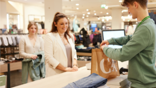 A C&A store employee assists a customer at the checkout counter, placing items into a C&A-branded shopping bag.