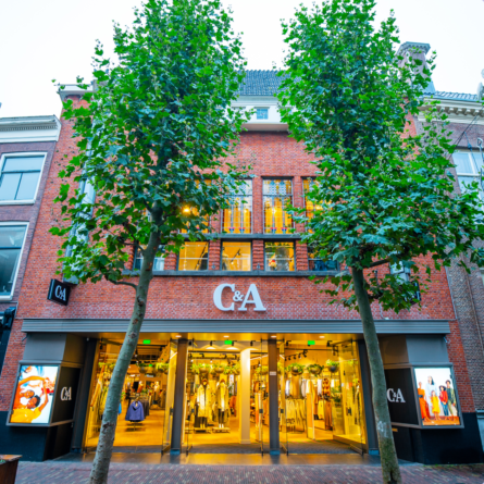 The exterior of a C&A retail store, framed by two tall trees, with a view of the inviting interior through large glass windows.