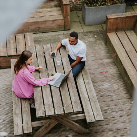 Two people, one in a pink jacket and the other in a white shirt, sitting at a wooden picnic table outside, working together on a laptop. The setting includes wooden benches and a paved area, with plants in the background.