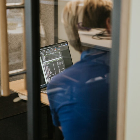 A person working intently on a laptop in a private office workspace, as seen through a glass partition.