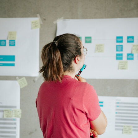 A woman in a red shirt thoughtfully looking at a wall covered with sticky notes and charts during a brainstorming session.