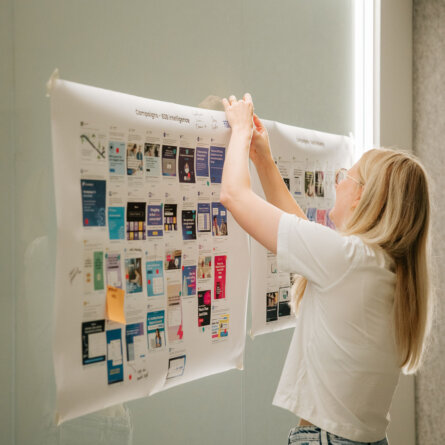 A woman in a white t-shirt attaching a large printout of campaign designs to a glass wall.