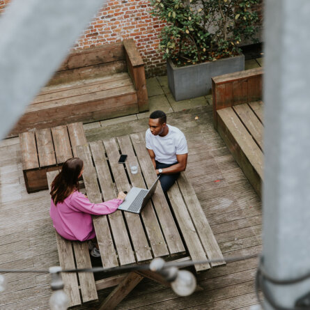 Overhead view of two colleagues discussing digital marketing strategies at a wooden picnic table in an outdoor setting surrounded by brick walls and greenery.