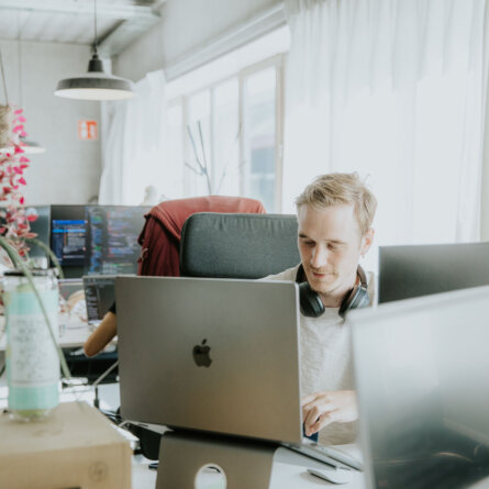 A developer working on a laptop in an open, plant-filled workspace with code displayed on screens in the background.