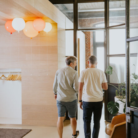 Two Colleagues Talking and Walking In a Modern Office, Wearing Light Colours And With Abstract Balloons in the Background