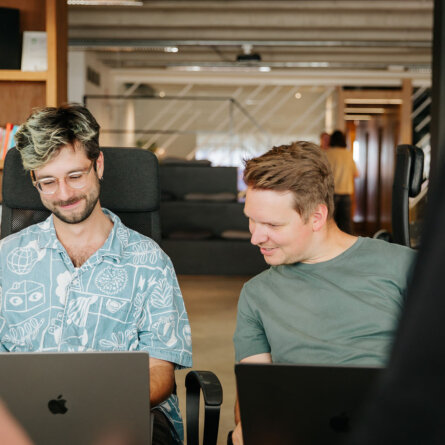 Two Colleagues Sitting Down And Discussing Something In a Cosy Office Looking at Macbook Screens, One Wearing A Blue Shirt and the Other Wearing a Dark Green Shirt